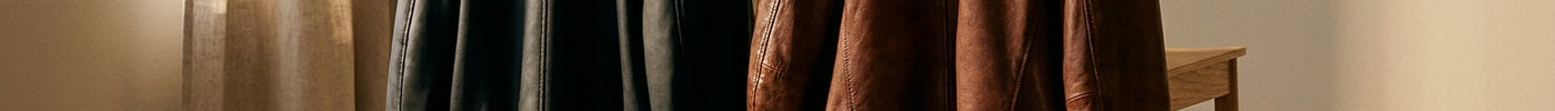 A faux leather jacket and a real leather jacket draped side by side over a wooden chair in a warmly lit, minimalist interior.
