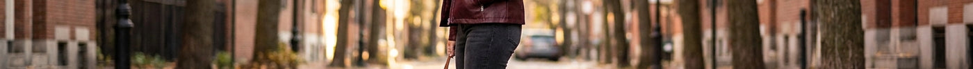 A stylish woman wearing a hooded faux leather jacket and dark jeans stands on an autumn street surrounded by golden falling leaves.